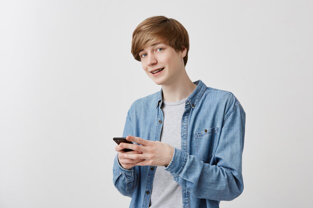 Modny mężczyzna o jasnych włosach i niebieskich oczach w dżinsowej koszuli pozuje w pomieszczeniu za pomocą telefonu komórkowego, rozmawia z przyjaciółmi, pisze wiadomość. Sprytny student wykorzystujący nowoczesne technologie, z uśmiechem