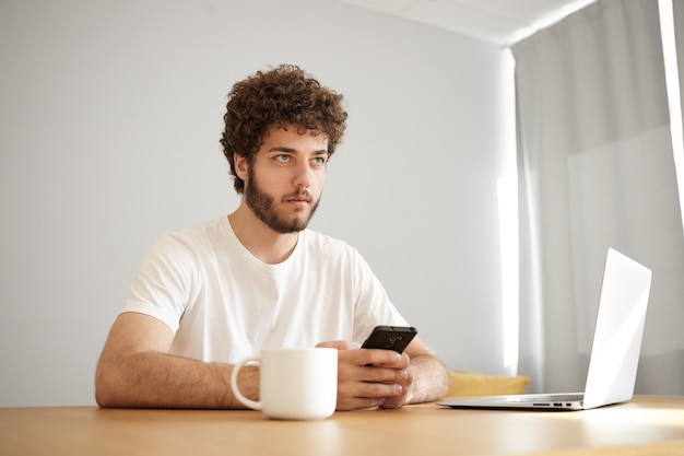 Modny młody, nieogolony facet z falującymi włosami i zamyślonym wyglądem pisze sms przez wifi na smartfonie, przegląda internet na przenośnym komputerze i pije kawę. Ludzie, styl życia i technologia
