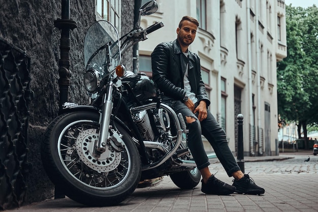 Modny motocyklista ubrany w czarną skórzaną kurtkę i dżinsy siedzący na swoim retro motocyklu na starej ulicy Europy.