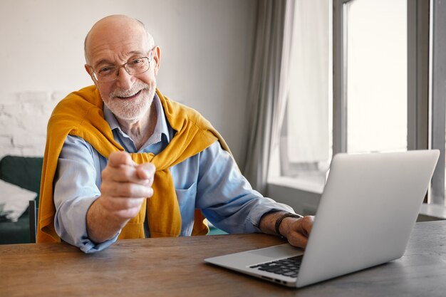 Modny, przyjazny starszy mężczyzna rekrutujący w prostokątnych okularach i eleganckich ubraniach, pracujący na przenośnym komputerze, uśmiechnięty szeroko i wskazujący palcem, wybierając Cię na stanowisko pracy