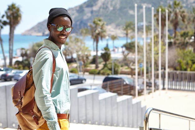 Modny student w kapeluszu i okularach przeciwsłonecznych, cieszący się słoneczną, ciepłą pogodą