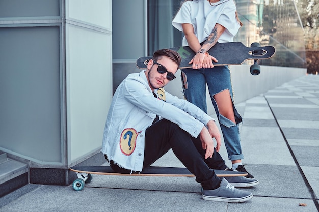 Modny ubrana para - młody hipster mężczyzna siedzi na longboard i jego dziewczyna stoi w pobliżu na zewnątrz.