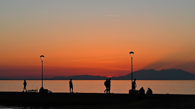 Molo z wieloma wędkarzami i spacerującymi ludźmi o zachodzie słońca, zaparkowany rower, latarnie lądowe, Grecja