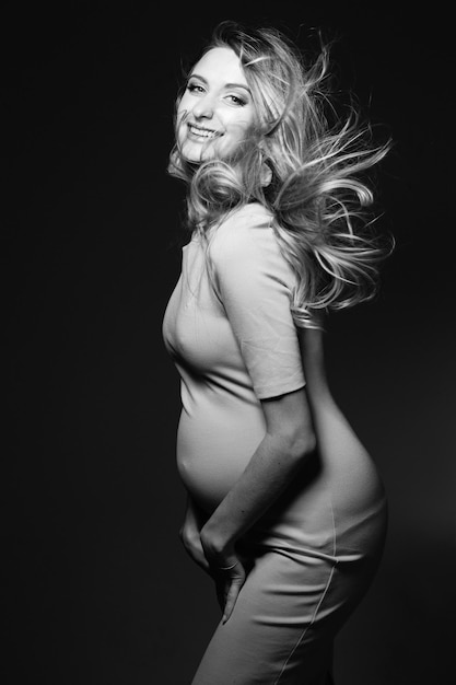 Monochromatyczny portret pięknej szczęśliwej kobiety w ciąży w sukni obejmującej brzuch i uśmiechnięty Pozytywność i stylowa kobieta pozowanie patrząc na kamery Koncepcja moda ciąża Windy effect