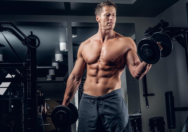 Muskularny mężczyzna shirtless robi biceps ćwiczenia z hantlami w klubie siłowni.