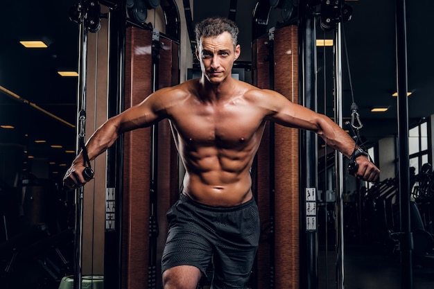 Muskularny mężczyzna shirtless robi ćwiczenia z aparatury treningowej w klubie ciemnej siłowni.