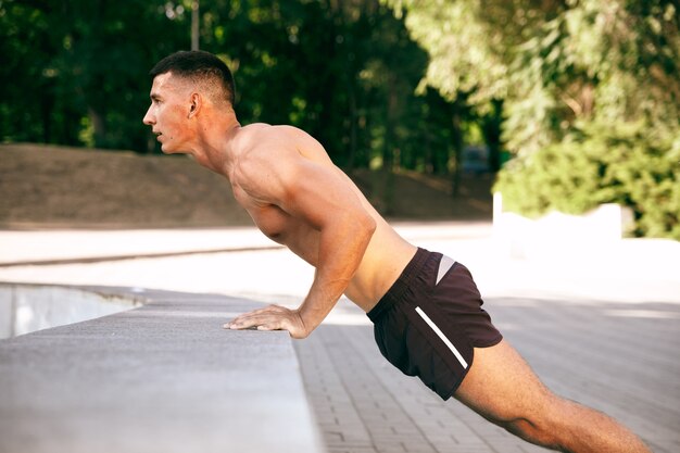 Muskularny mężczyzna sportowiec robi treningu w parku. Gimnastyka, trening, elastyczność treningu fitness. Letnie miasto w słoneczny dzień na tle pola. Aktywny i zdrowy tryb życia, młodość, kulturystyka.