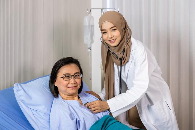 Muzułmańska kobieta lekarz za pomocą stetoskopu słuchać bicia serca pacjenta leżącego na łóżku i patrzącego na kamerę