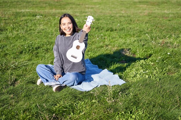 Bezpłatne zdjęcie muzyka i instrumenty portret słodkiej azjatyckiej dziewczyny pokazuje jej białe ukulele grające w parku podczas siedzenia