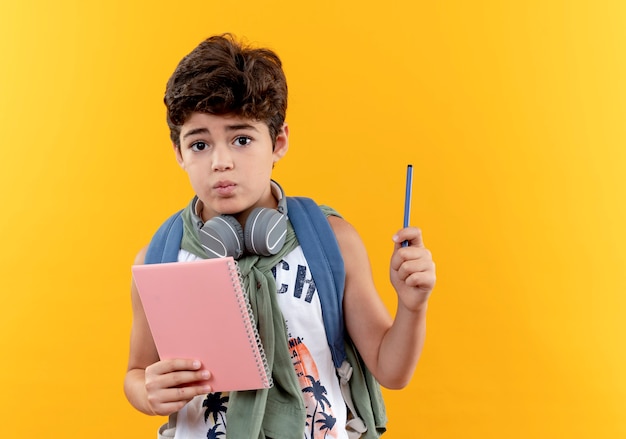 Mylić mały chłopiec w szkole noszenie plecaka i słuchawki trzymając notebook z piórem na białym tle na żółtym tle