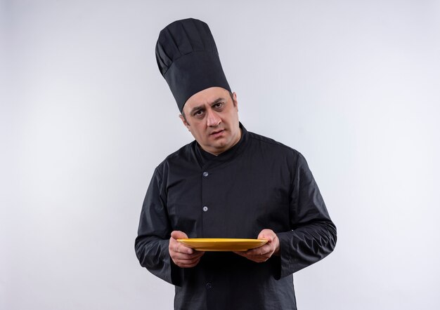 Mylić mężczyzna w średnim wieku kucharz w mundurze szefa kuchni trzymając talerz na na białym tle białej ścianie