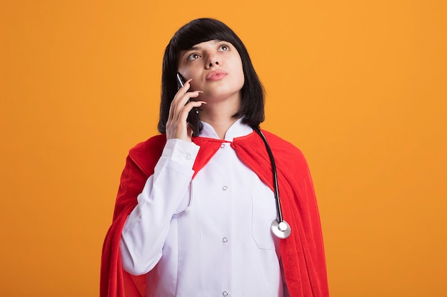 Myśląc, patrząc w górę młoda dziewczyna superbohatera w stetoskopie z szatą medyczną i płaszczem, mówi przez telefon odizolowany na pomarańczowej ścianie