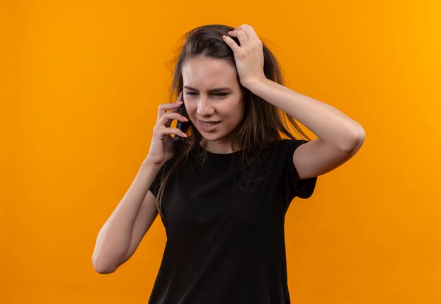 Myśląca młoda dziewczyna kaukaska ubrana w czarną koszulkę rozmawia przez telefon, kładzie rękę na głowie na odizolowanej pomarańczowej ścianie