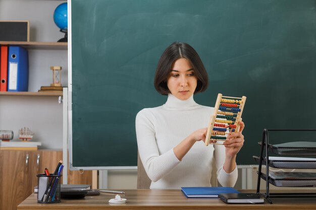 myśląca młoda nauczycielka trzymająca i patrząca na liczydło siedząca przy biurku z włączonymi narzędziami szkolnymi w klasie