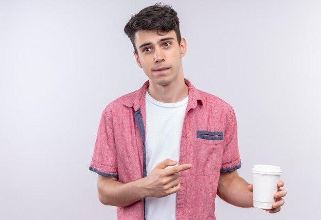 Myślący kaukaski młody facet ubrany w różową koszulę wskazuje na filiżankę kawy na na białym tle biały