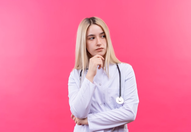 Myślący lekarz młoda blondynka ubrana w stetoskop w sukni medycznej położyła dłoń na brodzie na odosobnionej różowej ścianie