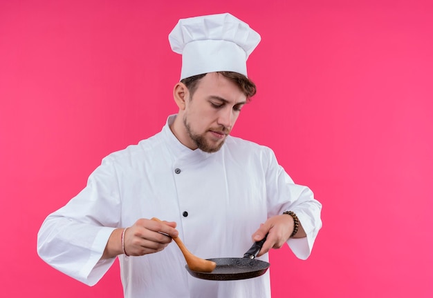 Myślący młody brodaty mężczyzna kucharz w białym mundurze trzyma patelnię z drewnianą łyżką, stojąc na różowej ścianie