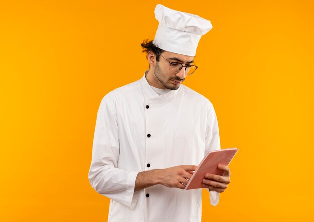Myślenie młody kucharz mężczyzna ubrany w mundur szefa kuchni i okulary, trzymając i patrząc na notebook na białym tle na żółtej ścianie