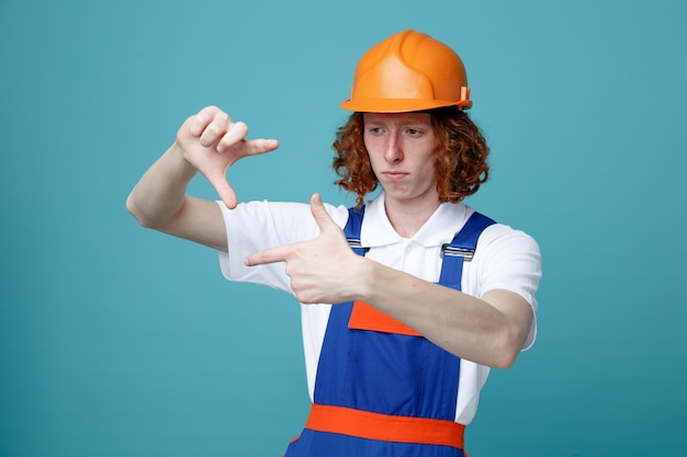 Myślenie pokazujący gest fotograficzny młody budowniczy mężczyzna w mundurze na białym tle na niebieskim tle