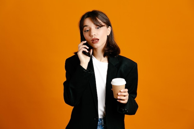 Myślenie trzymając filiżankę kawy rozmawia przez telefon młoda piękna kobieta ubrana w czarną kurtkę odizolowaną na pomarańczowym tle