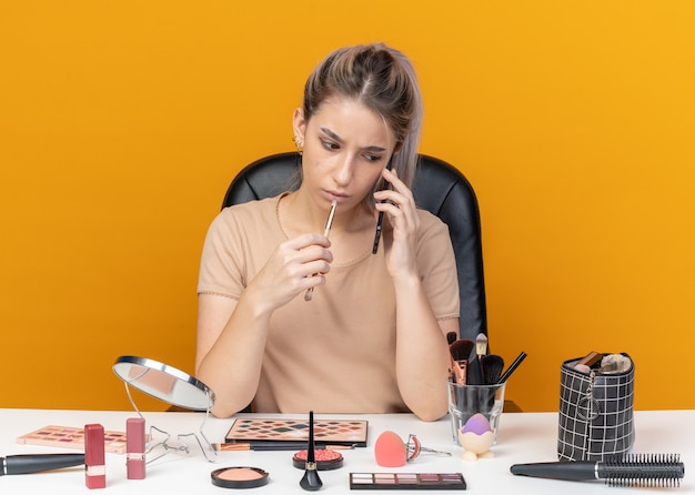 Myślenie, że młoda piękna dziewczyna siedzi przy stole z narzędziami do makijażu, trzymając pędzel do makijażu, mówi na telefonie na białym tle na pomarańczowym tle