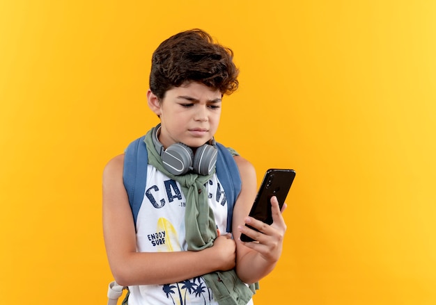Myśli mały chłopiec w szkole noszenie plecaka i słuchawek, trzymając i patrząc na telefon na białym tle na żółtej ścianie
