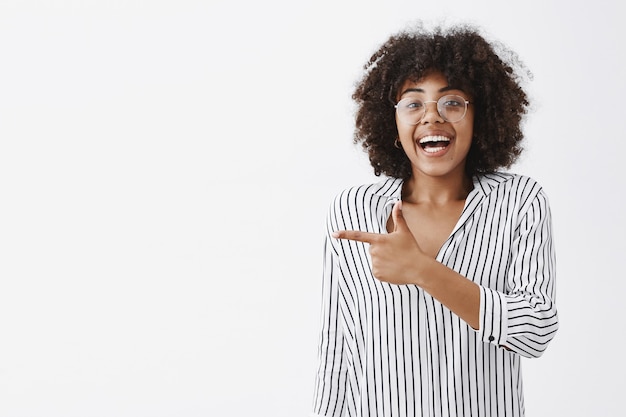 Nadmiernie emocjonalna, podekscytowana i szczęśliwa, urocza afroamerykańska kobieta w bluzce w paski i okularach, śmiejąca się z zabawy i radości, wskazująca palcem wskazującym w lewo, bawiąc się świetnie nad szarą ścianą