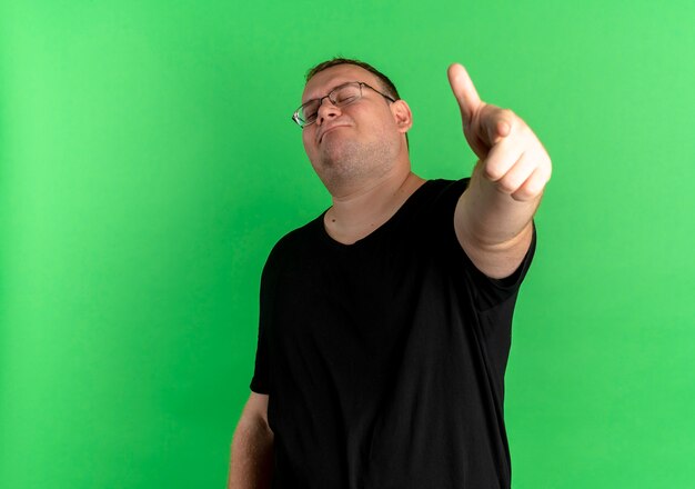 Nadwaga mężczyzna w okularach na sobie czarną koszulkę, pewny siebie, wskazując palcem wskazującym na zielonej ścianie