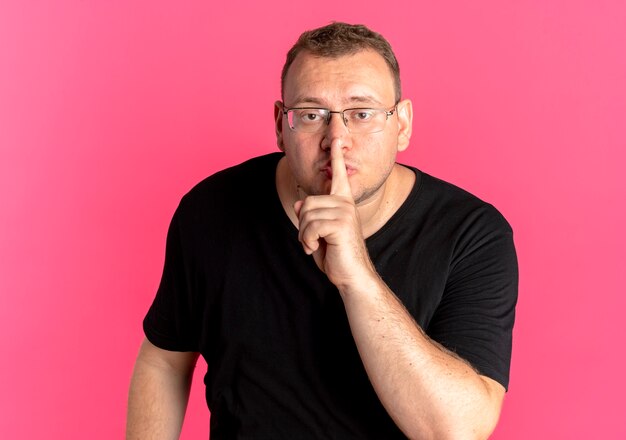 Nadwaga mężczyzna w okularach na sobie czarną koszulkę robi gest ciszy z palcem na ustach na różu