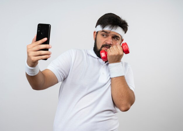 Napięty młody sportowy mężczyzna z opaską na głowę i nadgarstkiem, ćwiczenia z hantlami weź selfie na białej ścianie