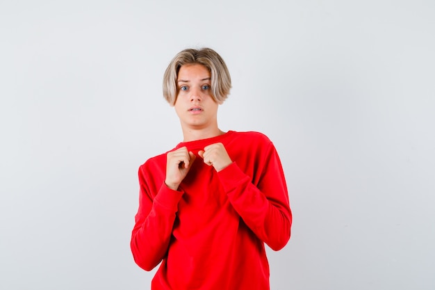 Nastolatek blond mężczyzna w czerwonym swetrze stojący w pozie walki i patrząc zakłopotany, widok z przodu.