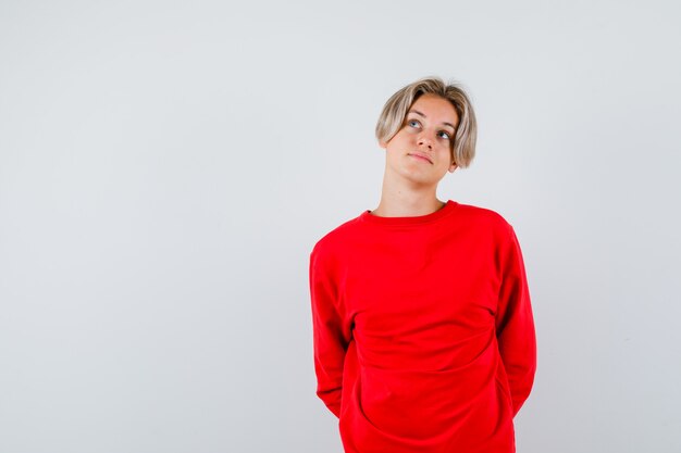 Nastoletni chłopiec trzymający ręce za plecami, patrzący w czerwony sweter i patrzący na zaabsorbowany. przedni widok.