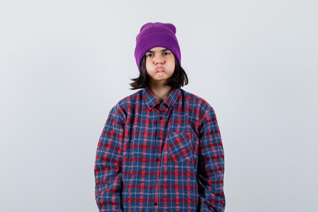 Nastoletnia kobieta wydmuchująca policzki w kraciastej koszuli i czapce wygląda na znudzoną