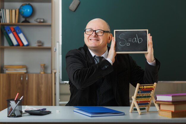 Nauczyciel w okularach siedzący przy ławce szkolnej przed tablicą w klasie pokazujący tablicę wyjaśniającą lekcję szczęśliwy i zadowolony, uśmiechający się radośnie