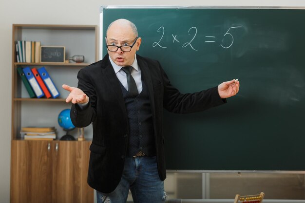 Nauczyciel w okularach stojący przy tablicy w klasie, wyjaśniający lekcję, patrząc zdezorientowany, podnosząc ręce z oburzeniem