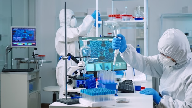 Naukowiec ubrany w kombinezon w wyposażonym laboratorium medycznym badający odkrycie leków za pomocą mikropipety. Biochemicy analizujący ewolucję wirusa za pomocą zaawansowanej technologicznie szczepionki badawczej przeciwko Covid19