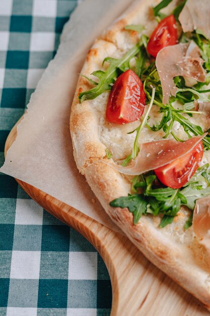 Neapolitańska pizza z szynką, serem, rukolą, bazylią, pomidorami posypanymi serem na drewnianej desce na obrusie w komórce z miejscem na tekst