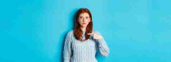Bezpłatne zdjęcie nerwowa i zdezorientowana ruda dziewczyna wskazująca na siebie stojącą w swetrze na niebieskim tle