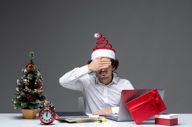 Nerwowy młody biznesmen z zabawnym czapką świętego mikołaja świętuje Boże Narodzenie i zamyka oczy w biurze na ciemnym tle