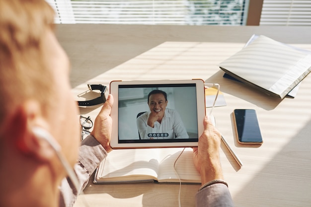 Bezpłatne zdjęcie nie do poznania mężczyzna siedzi przy biurku i rozmowy wideo na tablecie