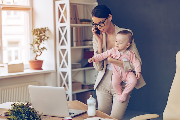 Nie jest łatwo być pracującą mamą! młoda piękna kobieta rozmawia przez telefon komórkowy i patrzy na laptopa, stojąc z córeczką w jej miejscu pracy