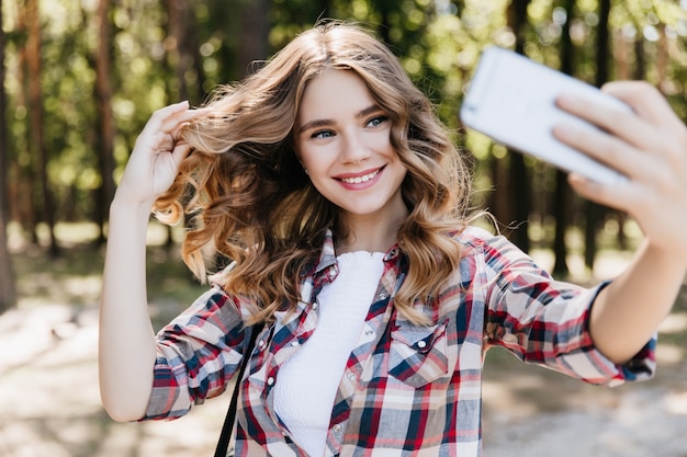 Niebieskooka nieśmiała dziewczyna przy użyciu telefonu do selfie w parku lato. Plenerowy portret eleganckiej blondynki pani bawi się włosami.