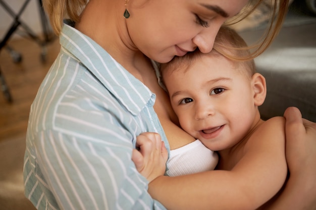 Niemowlęctwo, dzieciństwo i macierzyństwo. Kochająca piękna młoda matka przytulająca się w domu ze swoim uroczym uroczym synkiem, spędzająca szczęśliwe słodkie chwile, okazująca miłość i uczucie, uśmiechnięte dziecko