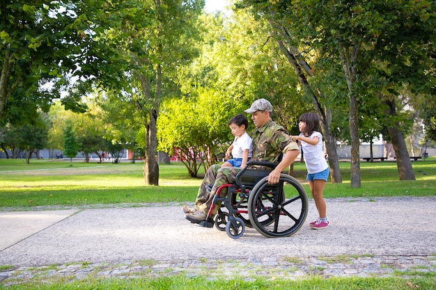 Niepełnosprawny weteran wojskowy spacerujący z dwójką dzieci w parku. Chłopiec siedzi na kolanach tatusiów, dziewczyna pcha wózek inwalidzki. Weteran wojny lub koncepcji niepełnosprawności