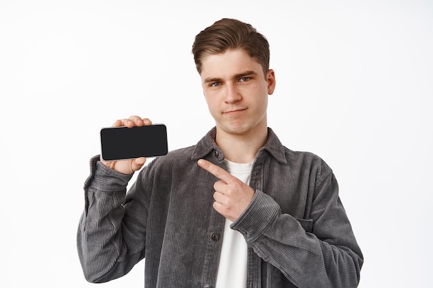Nierozbawiony młody mężczyzna wskazujący na poziomy ekran smartfona, pokazujący interfejs, aplikację zakupową lub grę mobilną, krzywiący się i marszczący brwi, sceptyczny, białe tło.