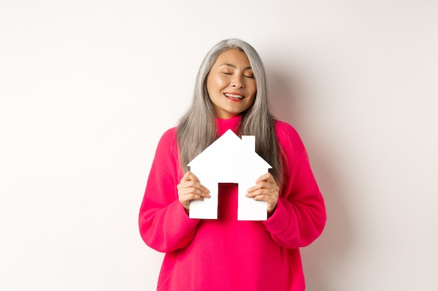 Nieruchomość. Piękna rozmarzona azjatycka dama przytulająca papierowy model domu z zamkniętymi oczami, uśmiechnięta jak marząca o kupnie mieszkania, stojąca na białym tle