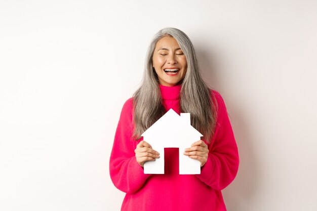 Nieruchomość. Szczęśliwa azjatycka babcia śmiejąca się z zamkniętymi oczami, trzymająca papierowy model domu, marząca o papierowym modelu domu, stojąca na białym tle