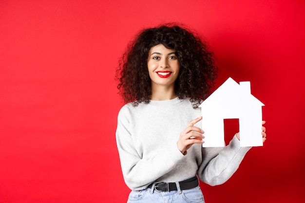 Nieruchomość. Uśmiechnięta kaukaski kobieta z kręconymi włosami i czerwonymi ustami, pokazująca papierowy model domu, szukająca nieruchomości, stojąca na czerwonym tle.