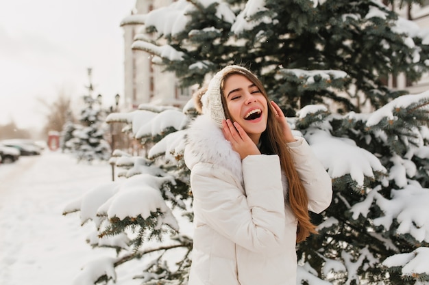 Niesamowita kobieta w białych ubraniach, zabawy w zimowy dzień, pozuje do zdjęcia. Zewnątrz portret zadowolony kaukaski kobieta na zaśnieżonej ulicy obok świerka.