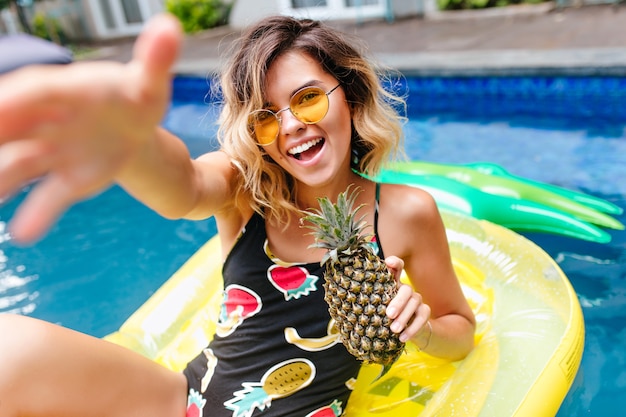 Niesamowita krótkowłosa dziewczyna w modnych okularach pływanie w basenie. Zewnątrz zdjęcie uroczej opalonej modelki w stylowym stroju kąpielowym, śmiejącej się w letni weekend.